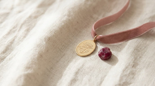 Goldener Anhänger mit gravierter Seerose neben einem tiefroten Rubin auf hellem Leinen