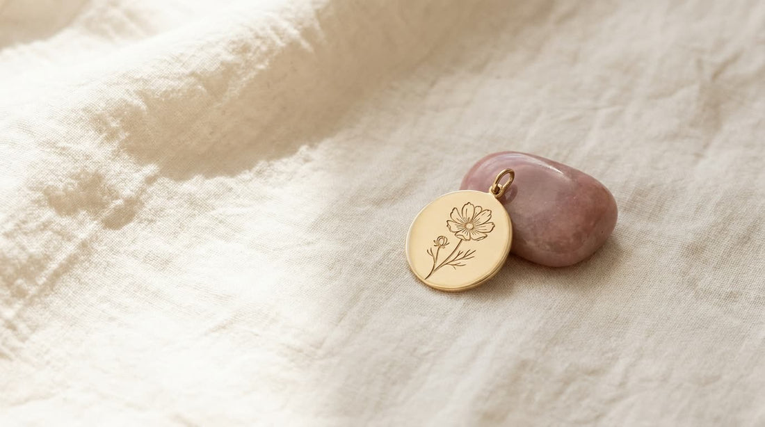 Goldener Anhänger mit gravierter Kosmee neben einem zart rosa Opal auf hellem Leinen